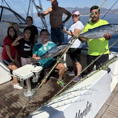 A young angler in the Blue Marlin cockpit fights a huge Bluefin Tuna, with another crew member assisting. Both are smiling and focused, with La Concha mountain and Marbella coast visible in the distance during a golden-hour Big Game fishing trip. Penn and Shimano reels are mounted on custom rods in holders. (Keywords: Big Game Fishing Marbella, Bluefin Tuna Catch, Blue Marlin Charter, Marina La Bajadilla Crew, Penn Shimano Gear)