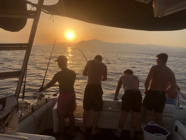Group of friends on a private sunset fishing charter in Marbella. Action shot with a bent rod during golden hour on the Blue Marlin boat, with the Mediterranean coast and mountains in the background.