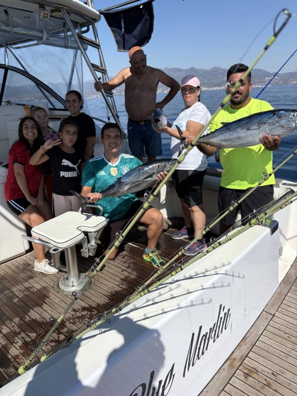 A young angler in the Blue Marlin cockpit fights a huge Bluefin Tuna, with another crew member assisting. Both are smiling and focused, with La Concha mountain and Marbella coast visible in the distance during a golden-hour Big Game fishing trip. Penn and Shimano reels are mounted on custom rods in holders. (Keywords: Big Game Fishing Marbella, Bluefin Tuna Catch, Blue Marlin Charter, Marina La Bajadilla Crew, Penn Shimano Gear)